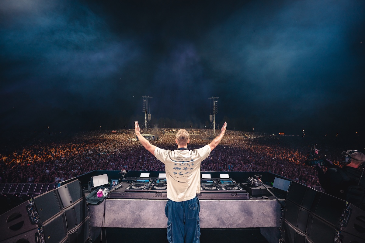 DJ Calvin Harris seen from behind with arms raised overlooking a massive outdoor festival crowd from the main stage decks during a live set.
